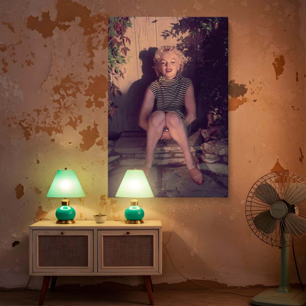 Marilyn Monroe appears thoughtful in this elegant vintage photo seated in a tucked-leg position on stone steps surrounded by foliage and rustic textures dressed in a sleeveless striped turtleneck ideal for wall art celebrating retro fashion and mid-centur