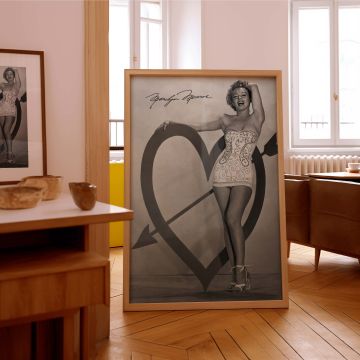 Stunning vintage glamour shot of Marilyn Monroe smiling brightly in a stylish and form-flattering outfit standing next to a heart arrow stage design with her hand on the heart and her signature above this print blends elegance fun and old Hollywood spirit